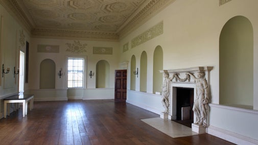 View of the Long Gallery in the house at Croome, with dark wooden floorboards and an ornately carved fireplace and ceiling